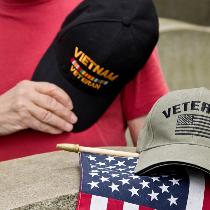 person wearing veteran baseball-style cap with head down over the top of a grave marker; a different cap and an American flag rest on top of the grave